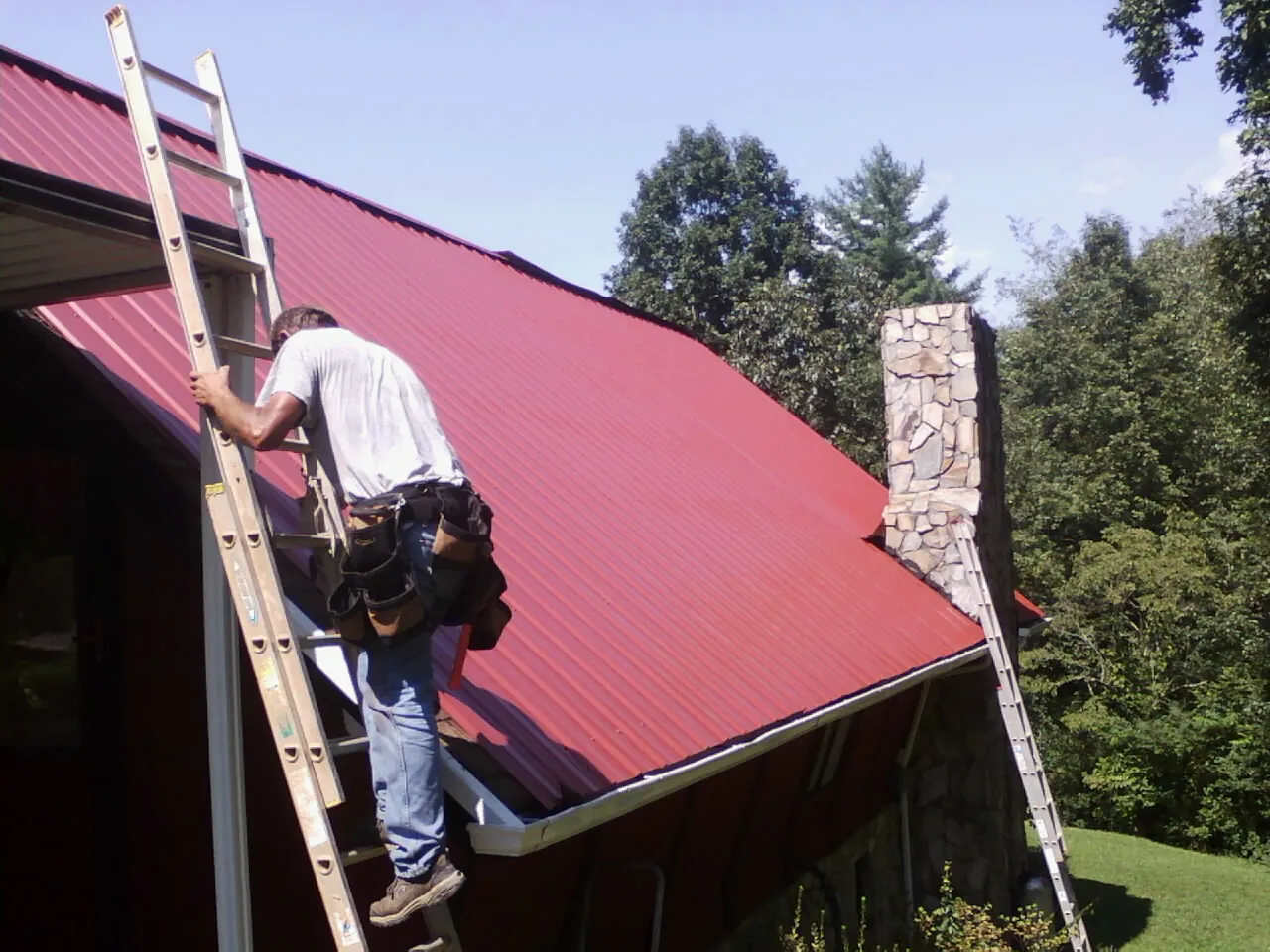 Roofer on a ladder beside a red metal roof and stone chimney — Soffit & Fascia Repair by Roofing Brothers in Brielle